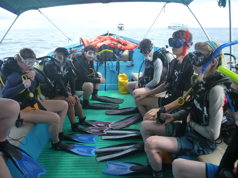 A group of people wearing scuba gear are sitting on a boat, preparing to go diving. They are surrounded by flippers and other diving equipment. The boat is on the water, and the sky is visible in the background. The divers appear excited and ready for their underwater adventure.